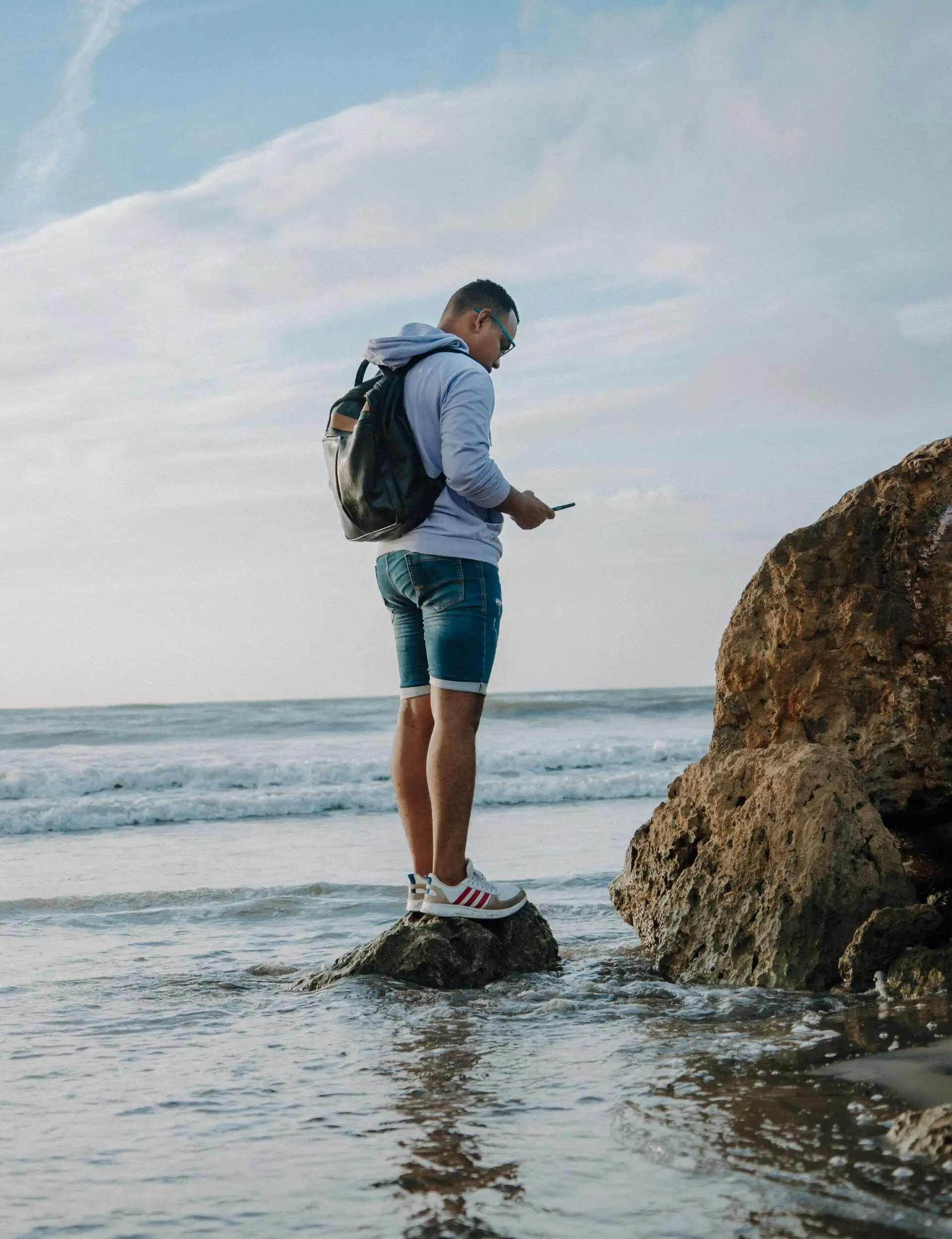 A man standing on a rock at the water's edge, looking at his phone